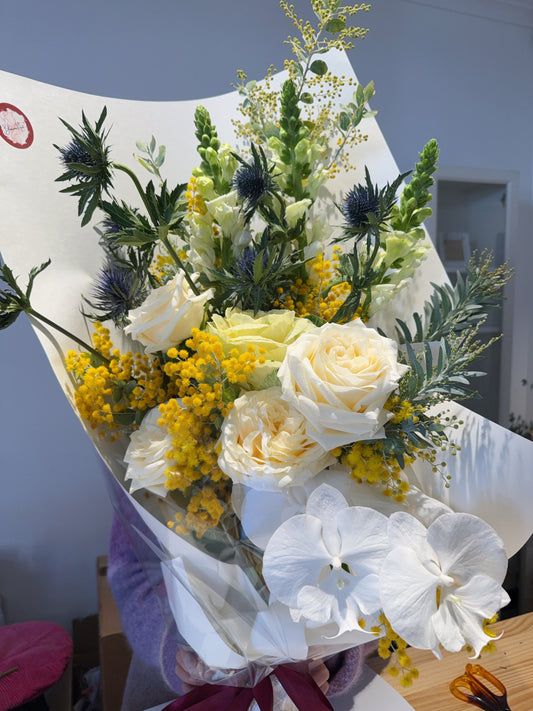 Bouquet of flowers with white roses, yellow wattle, and greenery on a white paper wrap.